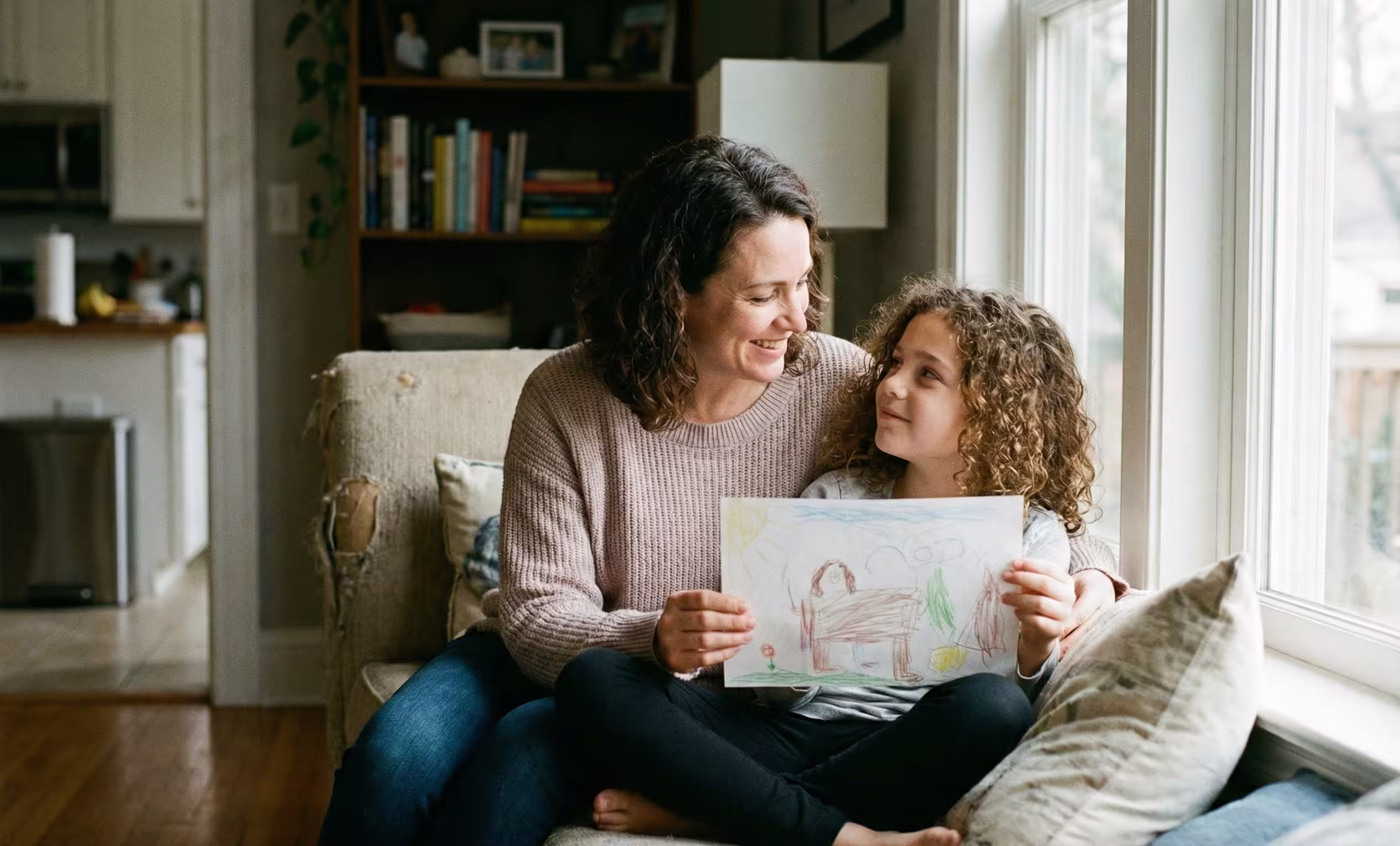 A mother shares a focused, intimate moment with her neurotypical child in a cozy window seat, illustrating the importance of seeing and supporting siblings of neurodiverse children, often called 'glass children.'