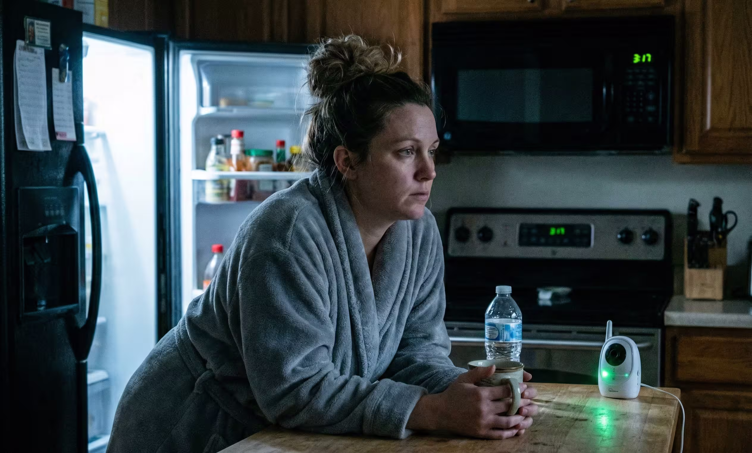 A tired mother in a robe stands in a dimly lit kitchen at 3:17 AM, holding a mug and staring blankly, illustrating the sleep deprivation experienced by parents of neurodiverse children.